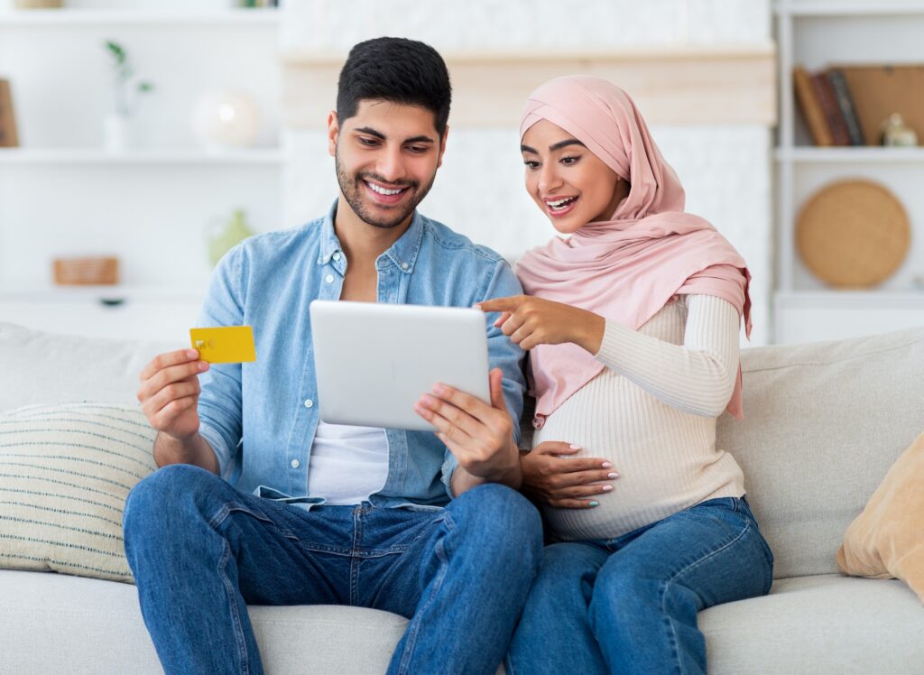Excited pregnant islamic spouses shopping online with tablet and credit card at home, relaxing